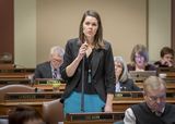 Representative Olson stands at her desk in the House Chamber and speaks into a microphone to someone out of view.
